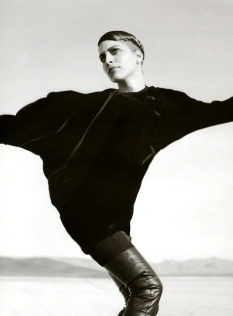 Black and white fashion shot of a slender adult woman in an oversized black blouse and leather boots posing in an open outdoor area.