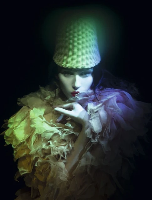 Fashion portrait of a woman wearing a basket hat and ruffled collar against a dark background, illuminated with blue and yellow light.