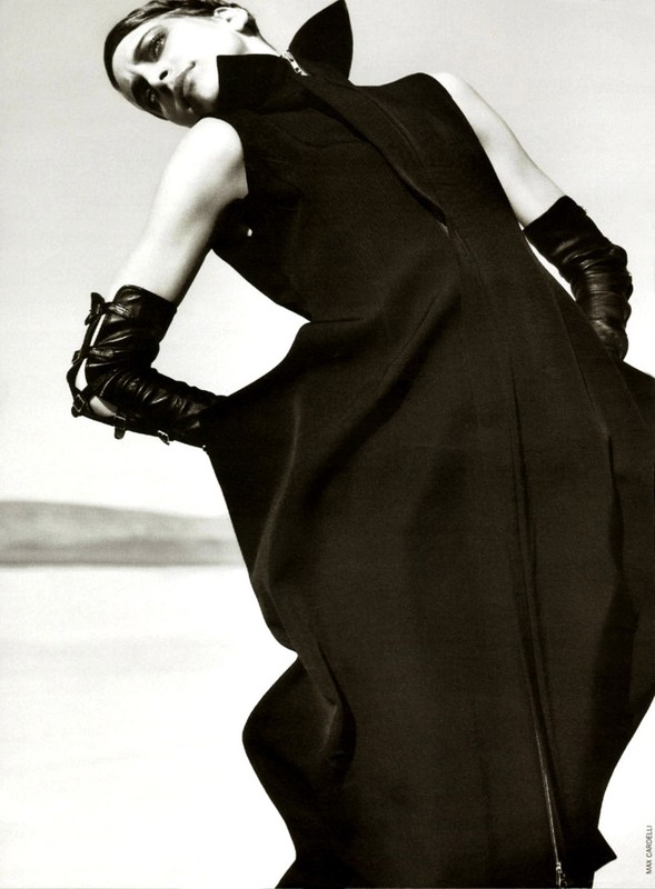 Model in a black high-collar dress and leather gloves posing against an outdoor horizon under a bright sky, photographed by Sarra Na.