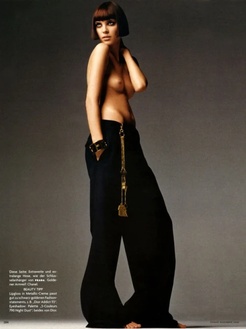 Fashion shot of a woman with a bob haircut, wearing black pants and a gold chain, posing in a studio environment with soft lighting.
