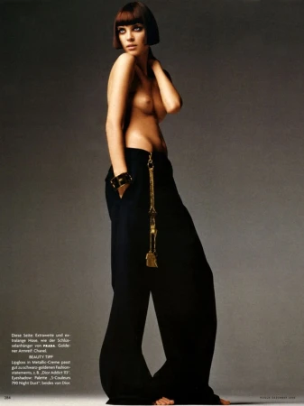 Fashion shot of a woman with a bob haircut, wearing black pants and a gold chain, posing in a studio environment with soft lighting.