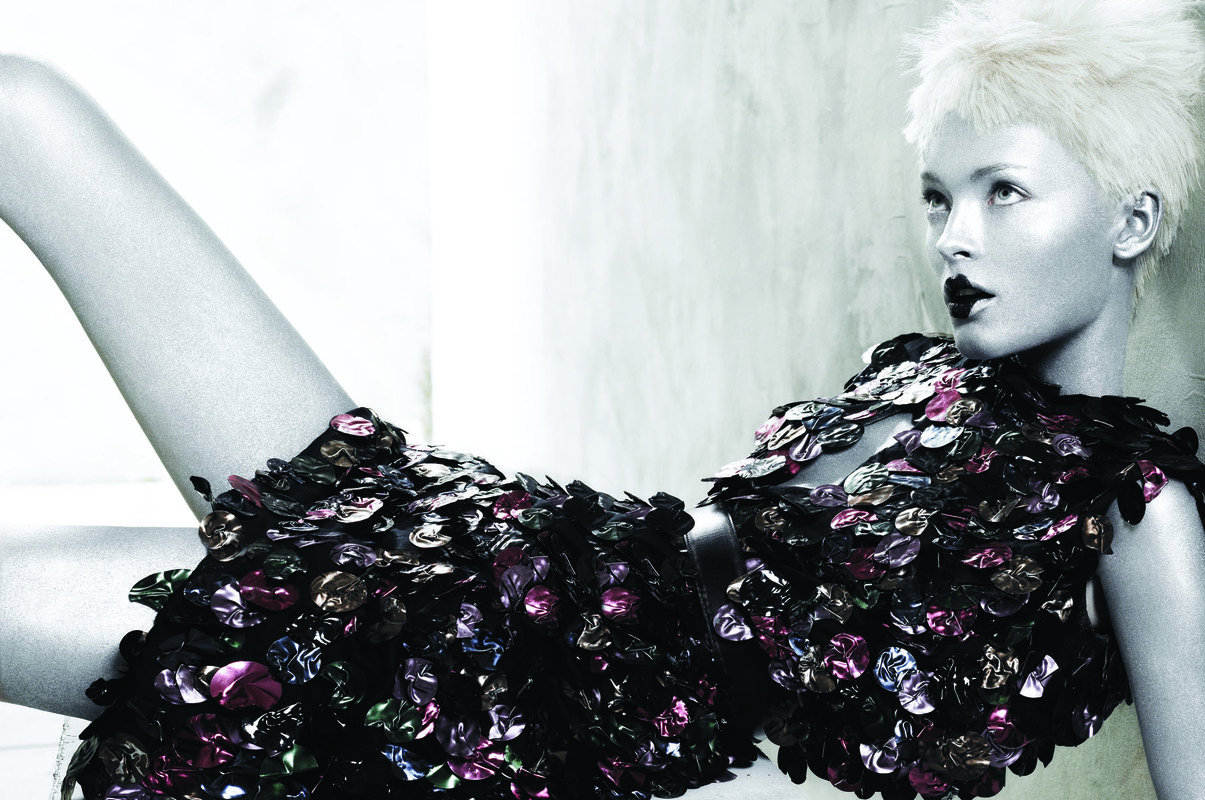 Fashion shot of a fair-skinned female model with platinum blonde hair wearing a dress made of glossy, dark, scale-like embellishments, studio setting.