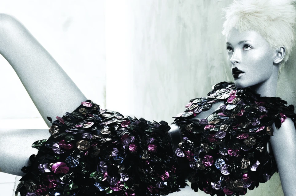 Fashion shot of a fair-skinned female model with platinum blonde hair wearing a dress made of glossy, dark, scale-like embellishments, studio setting.