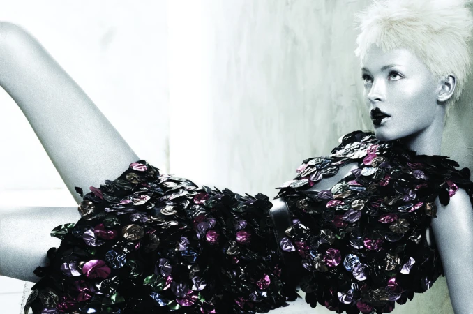 Fashion shot of a fair-skinned female model with platinum blonde hair wearing a dress made of glossy, dark, scale-like embellishments, studio setting.