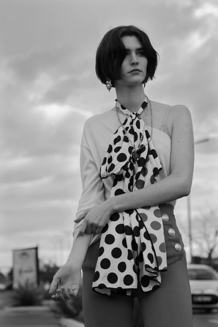 Black and white medium shot of a woman with short dark hair in a polka dot scarf, wearing an off the shoulder top outdoors on a cloudy day.