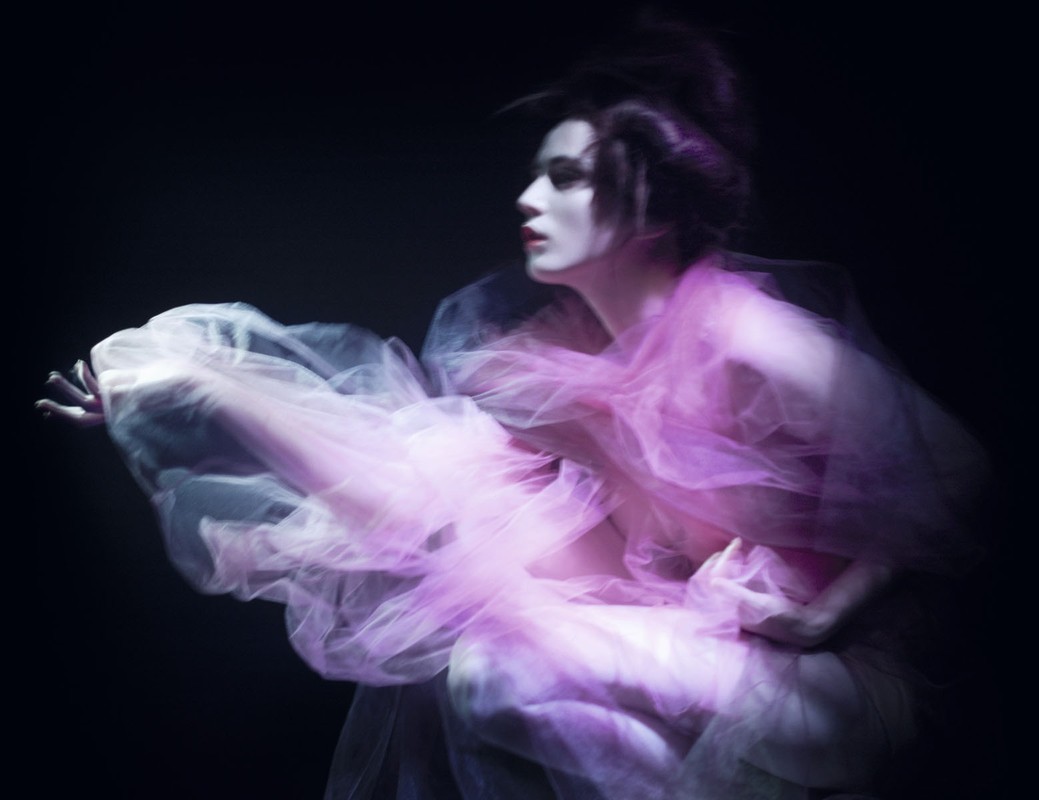 Side view of a person with dark hair, wearing a pink tulle dress, posed against a dark background in a fashion-style portrait.
