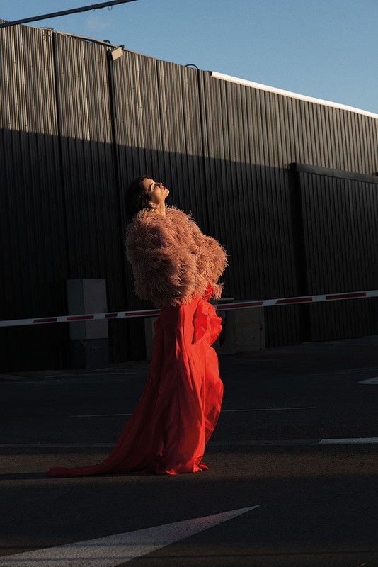 A woman in an orange dress and pink jacket stands in an open lot with a dark building in the background, looking up into the sunlight.