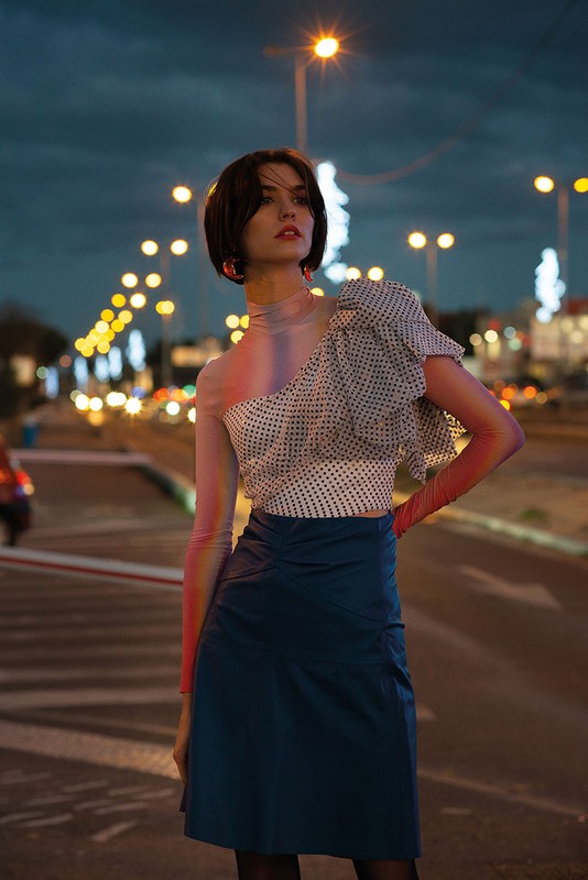 A woman in a polka dot blouse and blue skirt stands on a city street at night, with streetlights and traffic creating a blurred background.