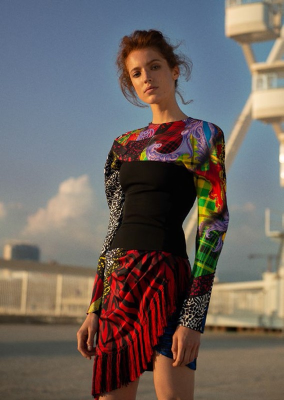 Young woman poses outdoors wearing a patchwork print dress against a daytime sky and structural background, captured in medium shot.