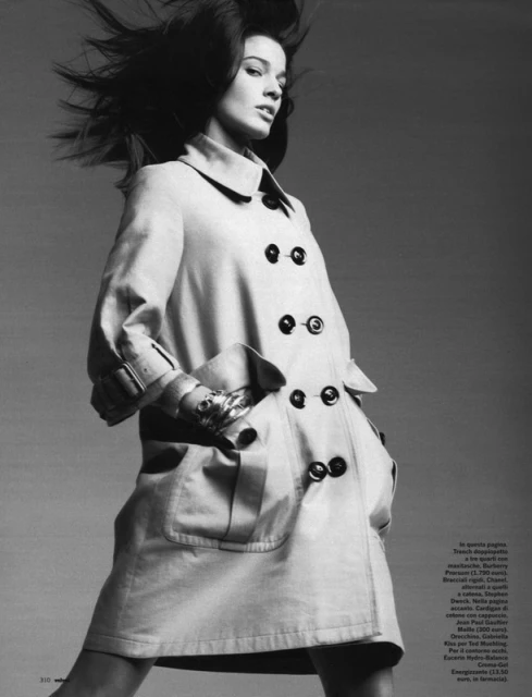 Black and white fashion shot of a woman in a trench coat, posing in front of a neutral background with wind-blown hair and jewelry.