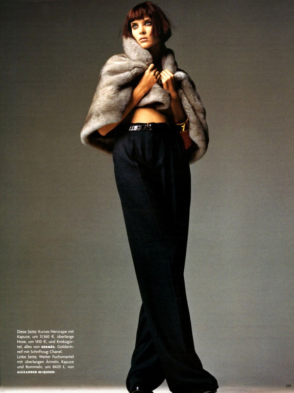 Fashion editorial shot of a woman in a fur cape, black pants, and a belt standing in a studio with a gray background and soft lighting.