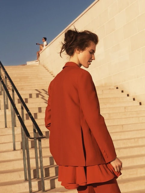 A woman in a red blazer and pants poses on stone stairs on a sunny day. Another person sits at the top of the stairs.