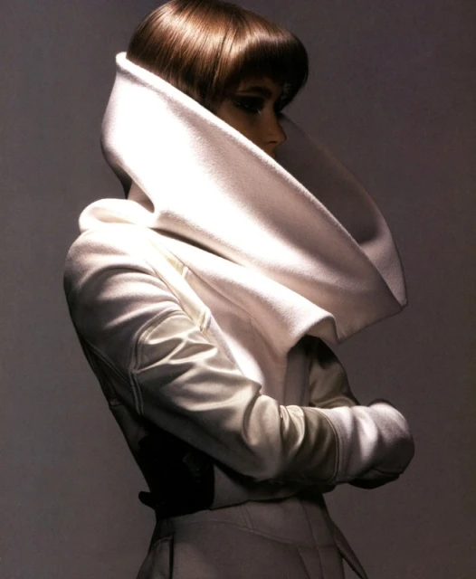 Woman with short brown hair poses in a studio wearing a structured white jacket with a large collar, side view, neutral backdrop.