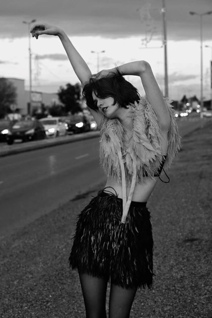 Black and white fashion shot of a woman in a feathered top and skirt standing on a road with cars and buildings in the background.