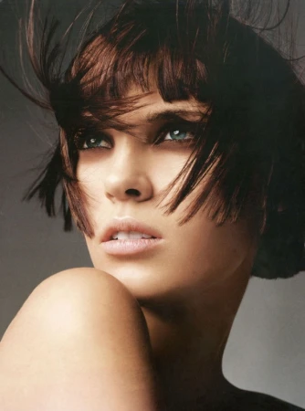 Close-up portrait of a woman with dark hair and striking eyes, with stray strands blowing across her face against a neutral background.