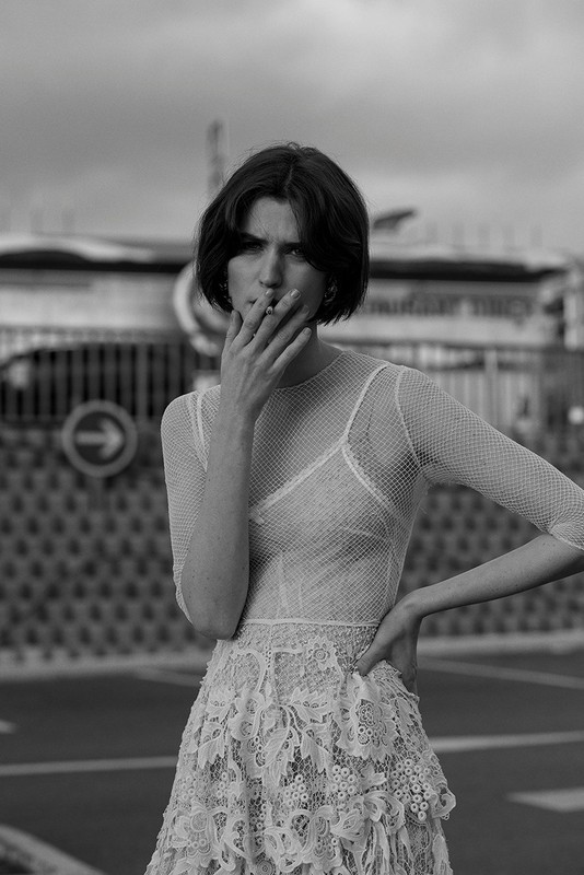Black and white portrait of a woman in a lace dress smoking a cigarette outdoors against a city street and building backdrop.