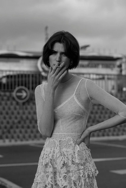 Black and white portrait of a woman in a lace dress smoking a cigarette outdoors against a city street and building backdrop.