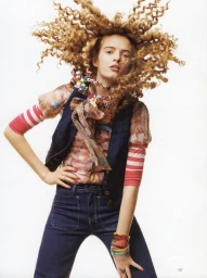 Woman with curly blonde hair flying outwards wearing dark vest over patterned top and blue jeans against white background