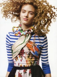 Portrait of a woman with voluminous blonde curly hair wearing a blue and white striped top and a colourful beaded necklace against a white background.