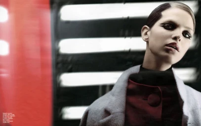 Model with dark eye makeup wearing a pale coat and red top against a striped background
