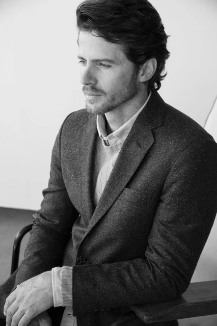 Black and white portrait of a man wearing a wool suit jacket and layered sweater sitting indoors looking away from the camera.