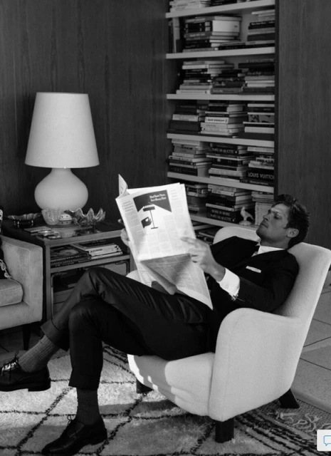Black and white portrait of a man reading a newspaper in an armchair, with a bookshelf and lamp visible in the background.