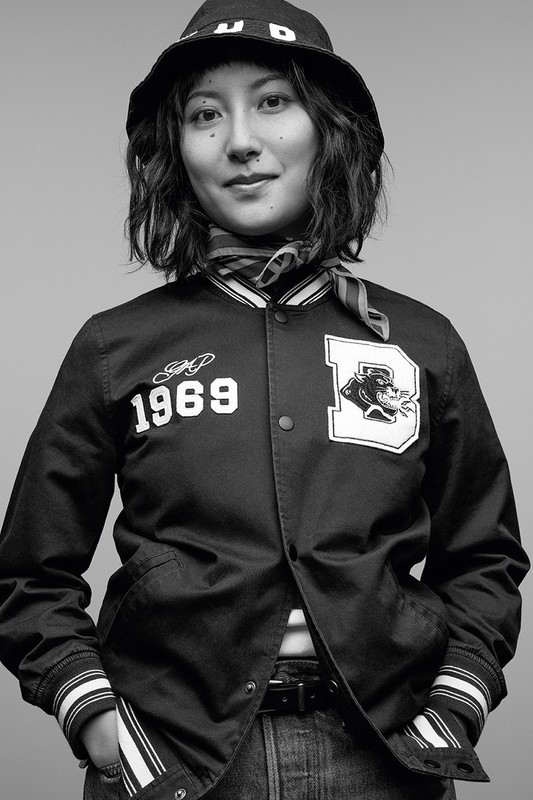 Black-and-white portrait of a young woman in a varsity jacket, jeans, and bucket hat, smiling slightly against a gray backdrop.