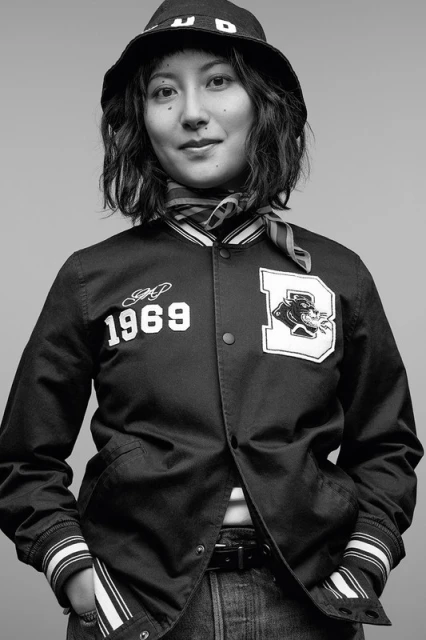 Black-and-white portrait of a young woman in a varsity jacket, jeans, and bucket hat, smiling slightly against a gray backdrop.