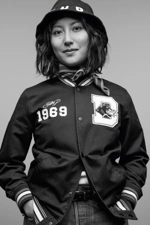 Black-and-white portrait of a young woman in a varsity jacket, jeans, and bucket hat, smiling slightly against a gray backdrop.