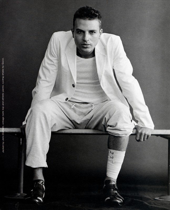 Monochrome portrait of a man in a white blazer and sweatpants sitting on a bench against a gray background in a studio environment.