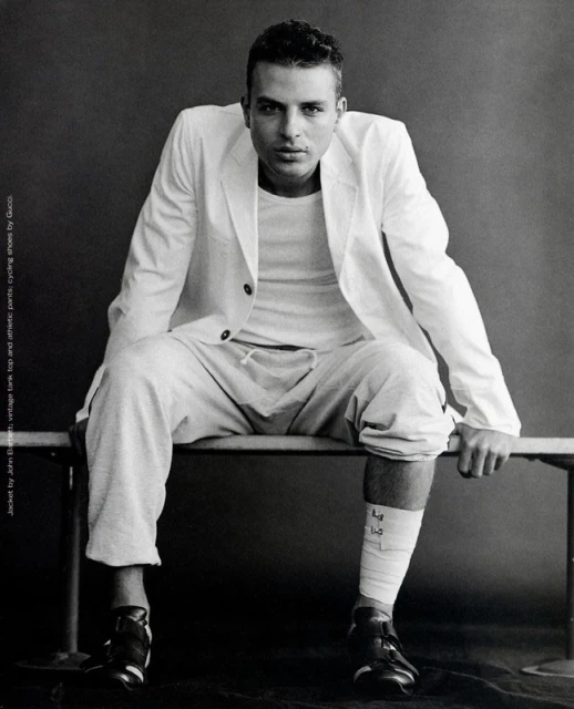 Monochrome portrait of a man in a white blazer and sweatpants sitting on a bench against a gray background in a studio environment.