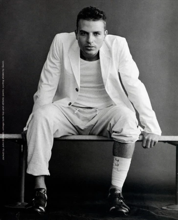 Monochrome portrait of a man in a white blazer and sweatpants sitting on a bench against a gray background in a studio environment.