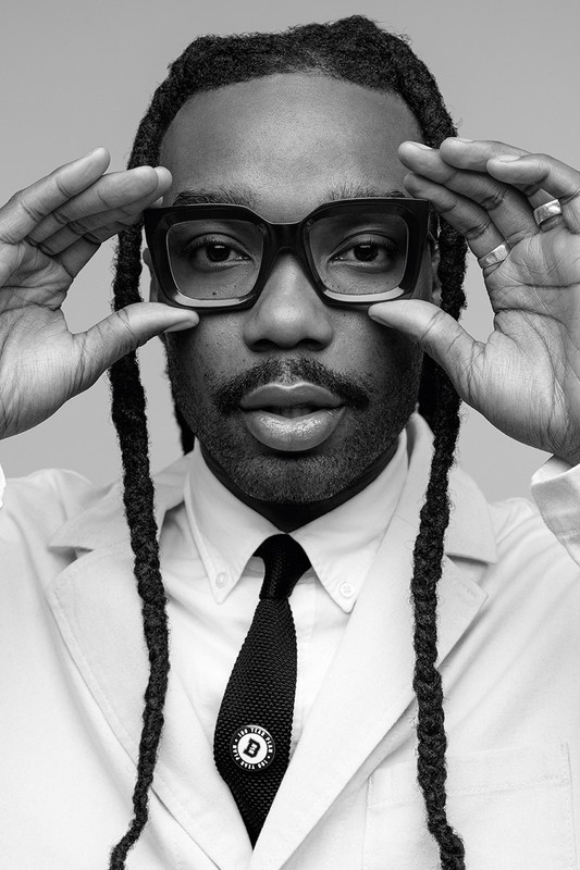 Monochrome portrait of a Black male in large glasses, wearing a lab coat and tie, looking at the camera with hands framing his face.