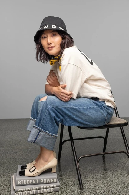 Young adult female poses seated indoors, wearing casual clothes, bucket hat, and pumps on stacked books in a studio environment.