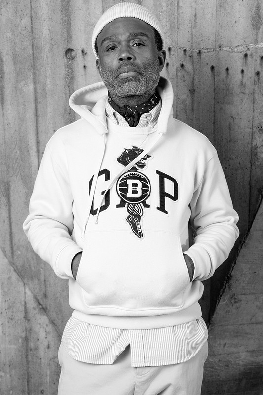 Portrait of an adult male with a beanie, wearing a graphic hoodie, standing indoors against a textured wall in a black and white photograph.