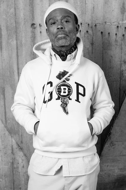 Portrait of an adult male with a beanie, wearing a graphic hoodie, standing indoors against a textured wall in a black and white photograph.