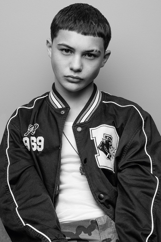 Close-up portrait of a young male model wearing a dark bomber jacket with white piping over a white tank top.