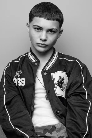 Close-up portrait of a young male model wearing a dark bomber jacket with white piping over a white tank top.