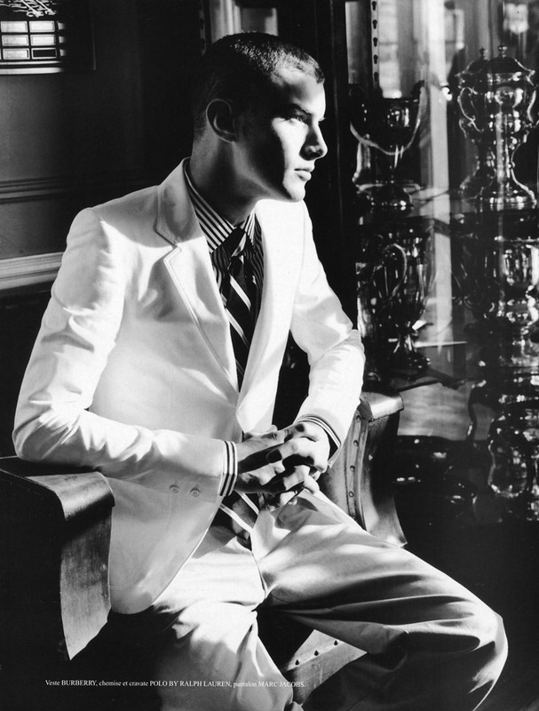 Black and white fashion shot of a man in a white suit sitting in a chair, looking away from the camera with his hands clasped together.