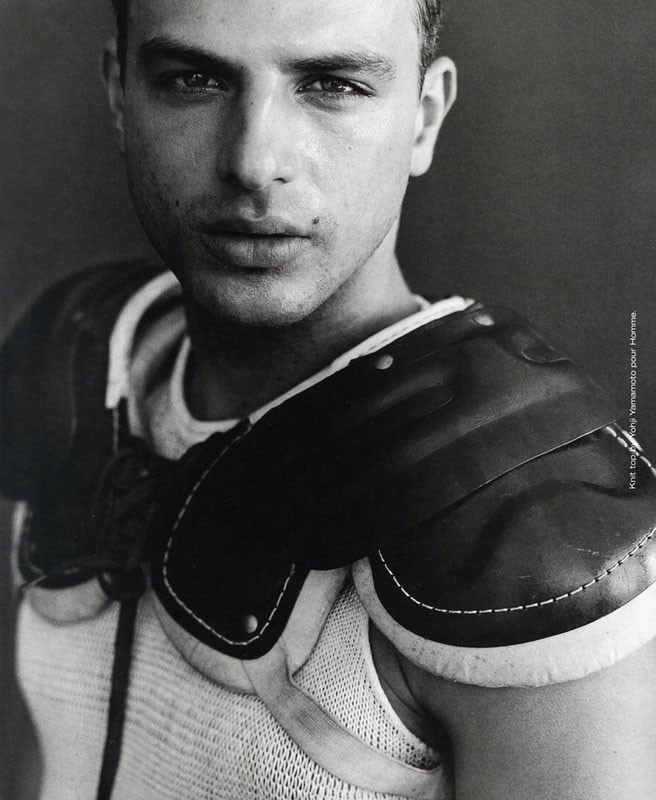 Monochromatic portrait of a male model wearing shoulder pads and a knit top against a dark background in an indoor setting.