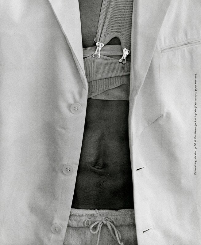 Close-up studio shot of a person with an open white linen jacket, neutral-colored knit top with decorative bone-shaped clasps, and light grey sweatpants.
