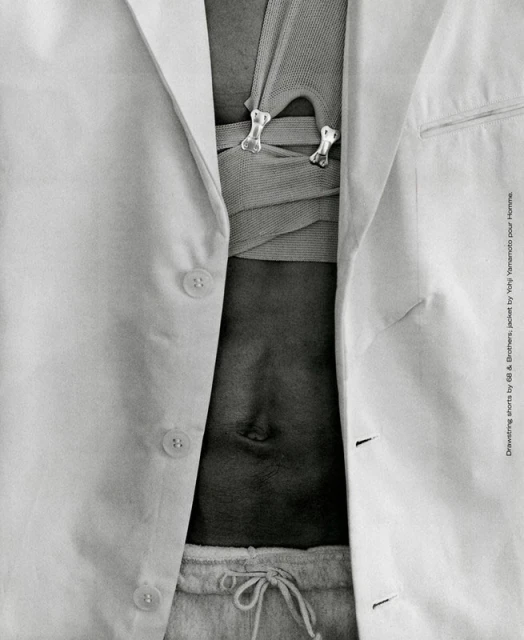 Close-up studio shot of a person with an open white linen jacket, neutral-colored knit top with decorative bone-shaped clasps, and light grey sweatpants.