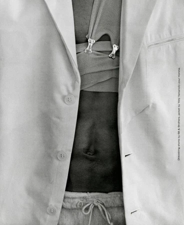 Close-up studio shot of a person with an open white linen jacket, neutral-colored knit top with decorative bone-shaped clasps, and light grey sweatpants.
