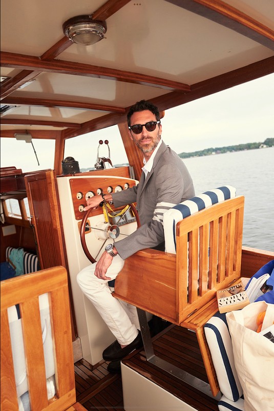 Man piloting a wooden boat on the water wearing a grey suit jacket, white pants, and sunglasses on a sunny day.