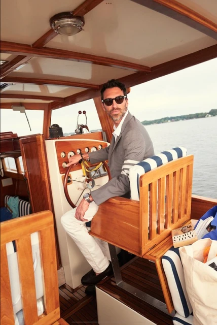 Man piloting a wooden boat on the water wearing a grey suit jacket, white pants, and sunglasses on a sunny day.