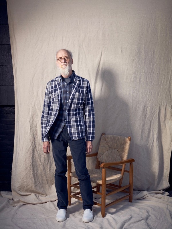 Full-length portrait of a senior man in a blue plaid jacket and black pants, standing next to a wooden chair against a neutral backdrop.