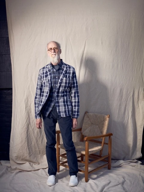 Full-length portrait of a senior man in a blue plaid jacket and black pants, standing next to a wooden chair against a neutral backdrop.