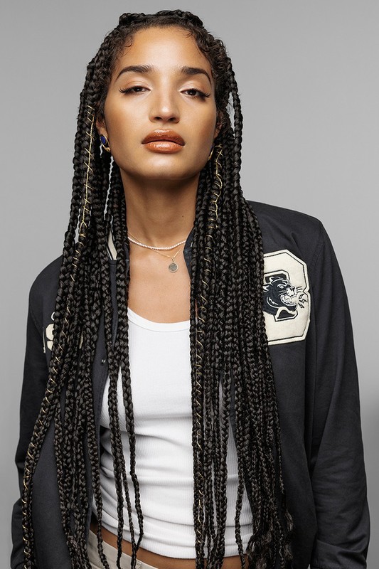 Portrait of a woman with long braided hair wearing a dark varsity jacket and white tank top against a grey background.