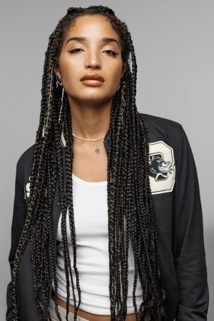 Portrait of a woman with long braided hair wearing a dark varsity jacket and white tank top against a grey background.