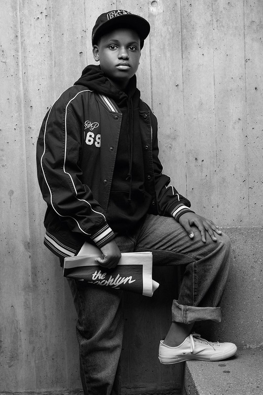 Black boy in a cap and varsity jacket holding a folded newspaper against a textured concrete wall